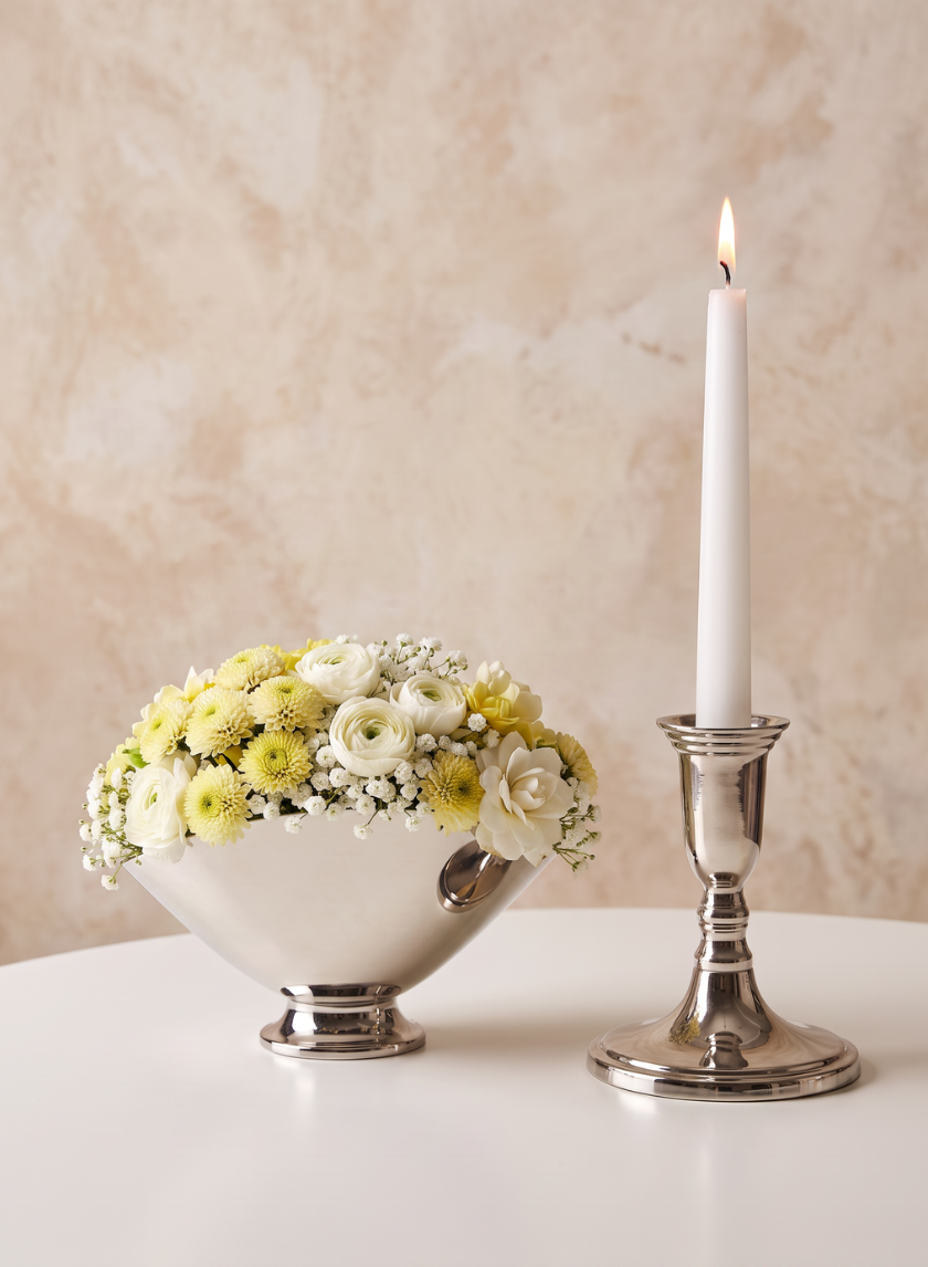 A silver vase with white and yellow flowers sits on a round table next to a lit white candle in the 5.5in Tall Nickel Silver Candlestick, against a beige textured background.