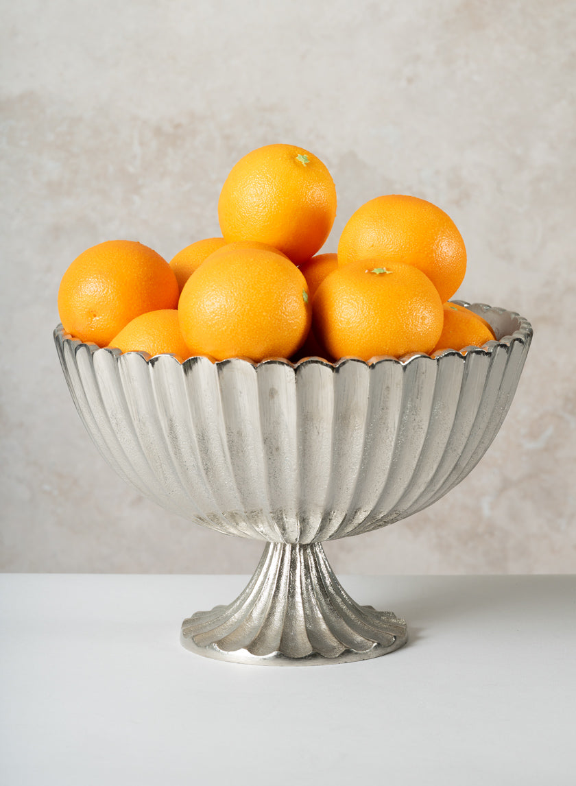 A 12in Palermo Brushed Silver Scallop Compote Metal Bowl filled with whole oranges sits on a white surface against a light, textured background.