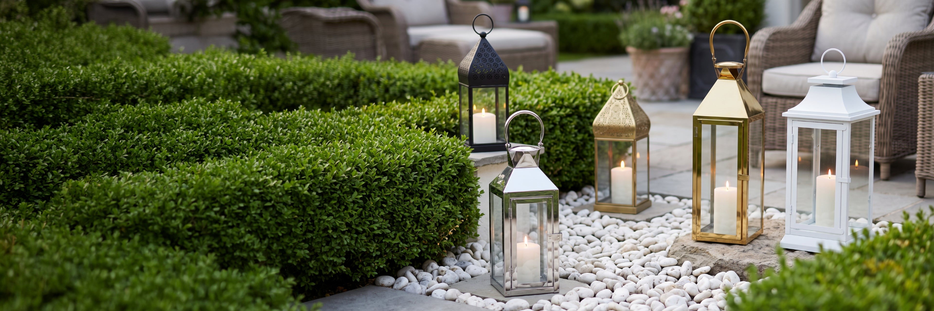Four decorative lanterns in black, gold, and white finishes with lit candles inside, arranged on white pebbles next to a neatly trimmed green hedge in an outdoor patio setting.
