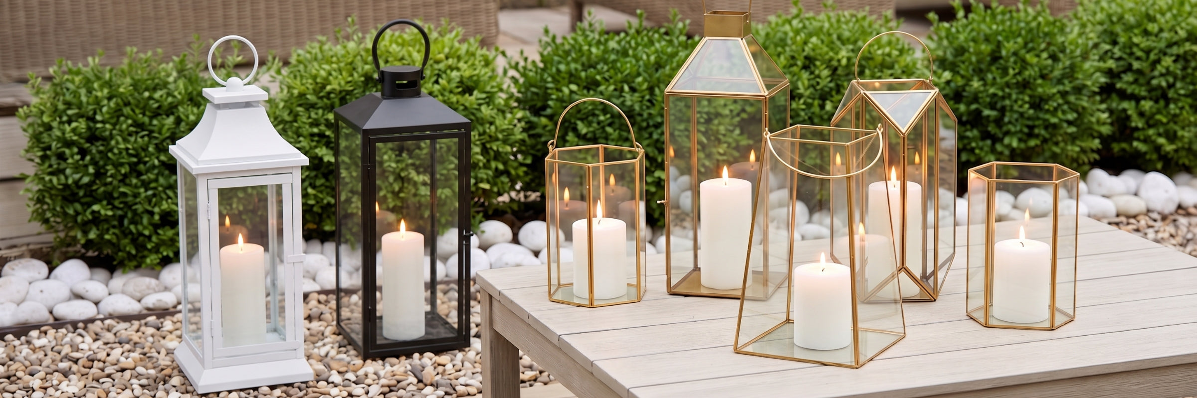 A collection of six glass lanterns and candle holders with gold and black metal frames, filled with white pillar candles and lit, arranged on a light-colored wooden table outdoors in front of green shrubs.