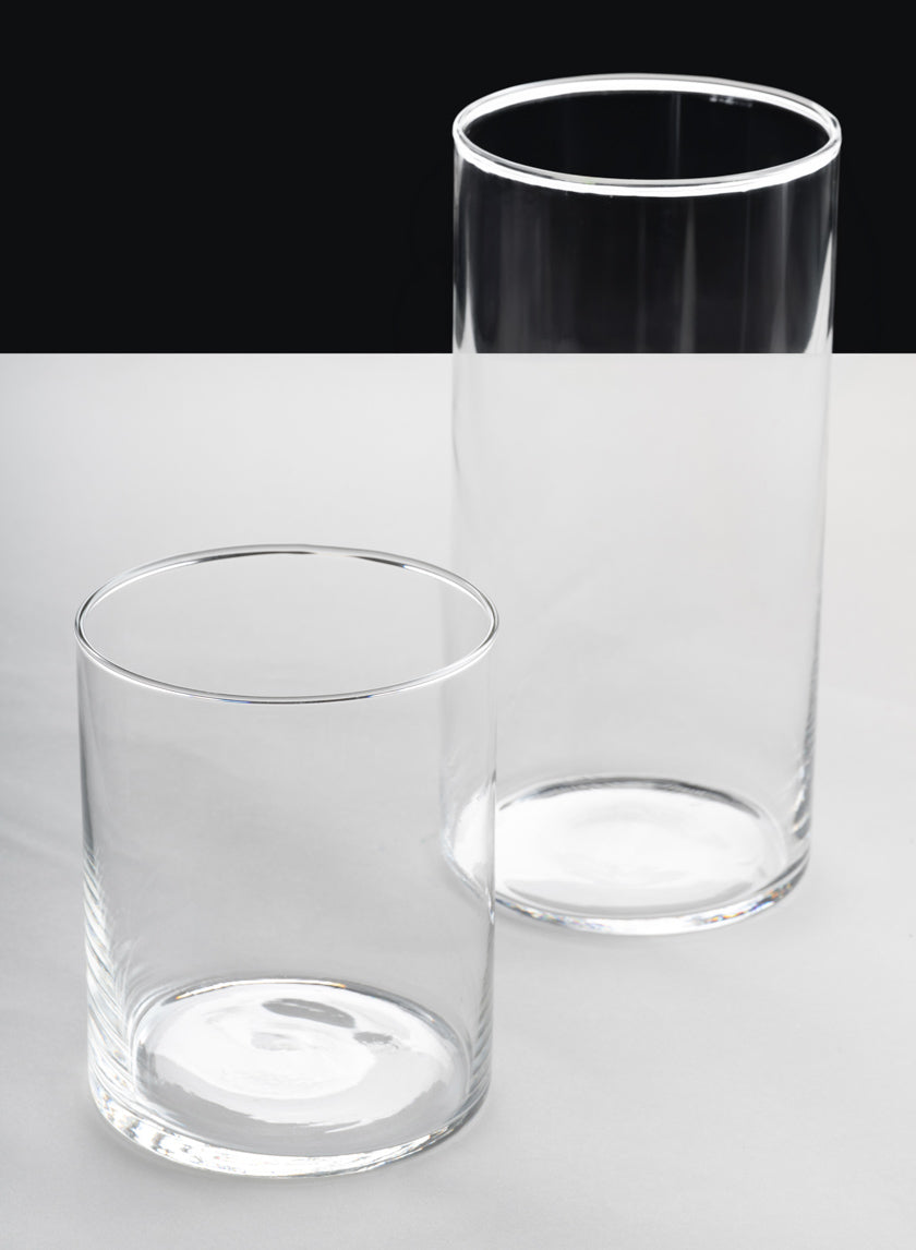 Two empty Best Value Glass Cylinder Vases (Set of 6) are side by side on a light surface, one taller and set slightly behind the shorter. The background is two-toned: black on top, light gray below.