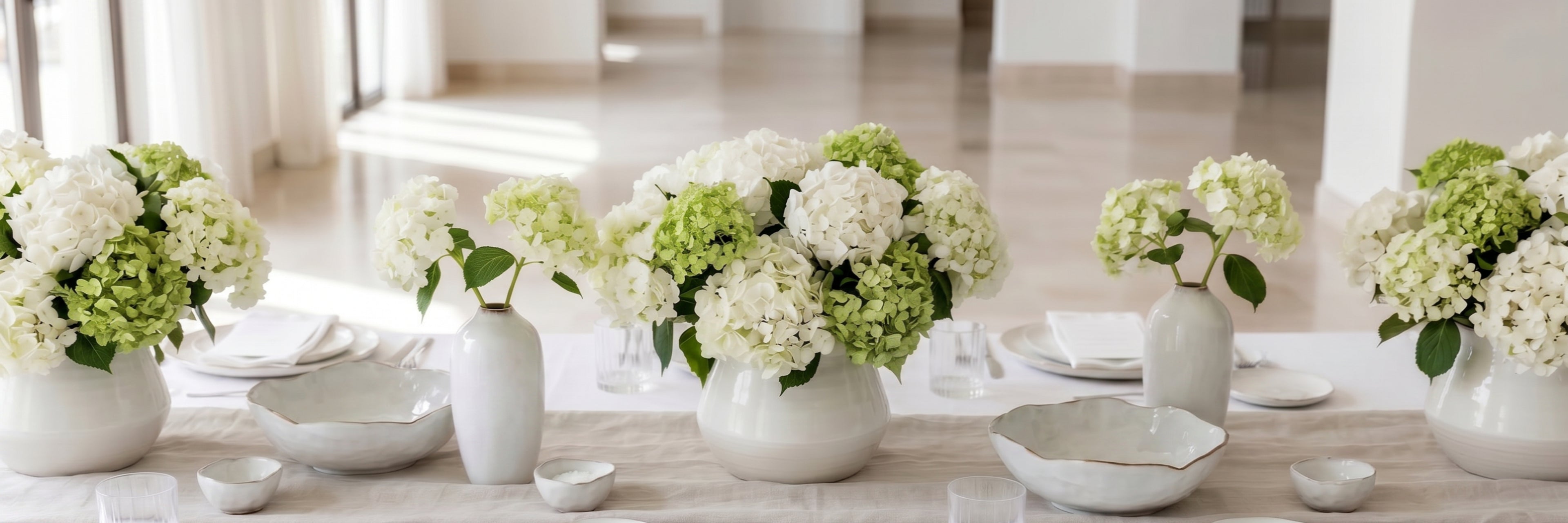 A long dining table set with white ceramic vases holding large white and light green hydrangea bouquets, alongside white plates and bowls in a bright, modern interior.