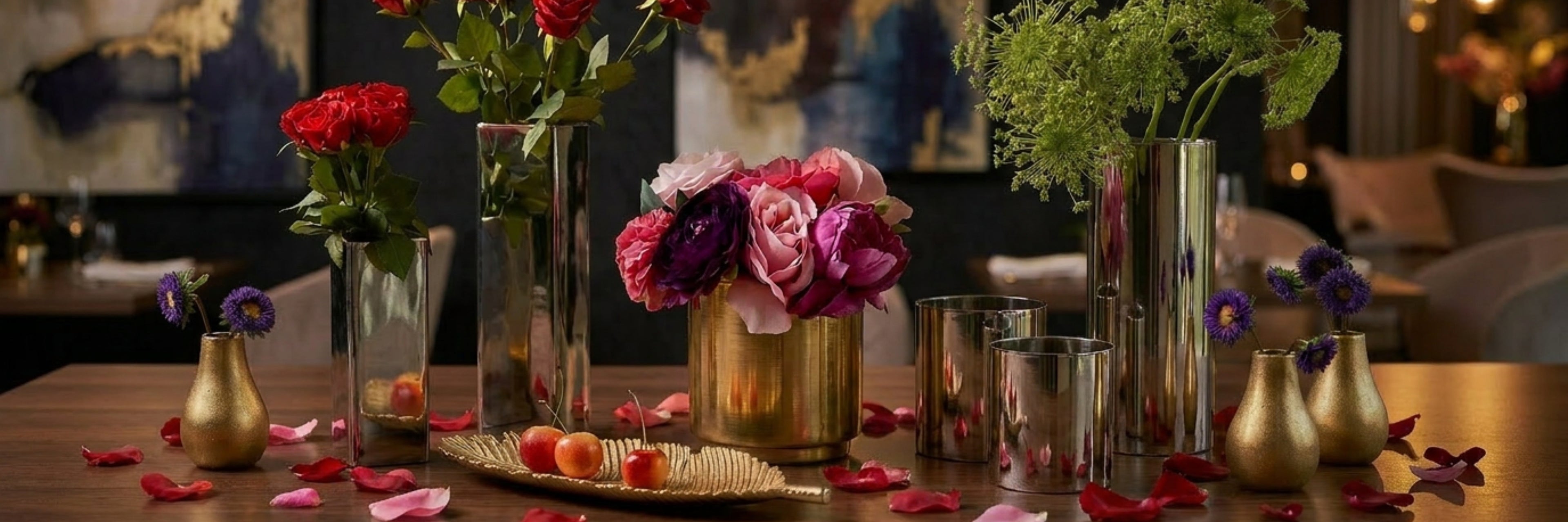 A styled tabletop arrangement of reflective metallic flower vases and small gold vases holding pink, red, purple, and green florals, with scattered rose petals on a wooden surface in an indoor setting.