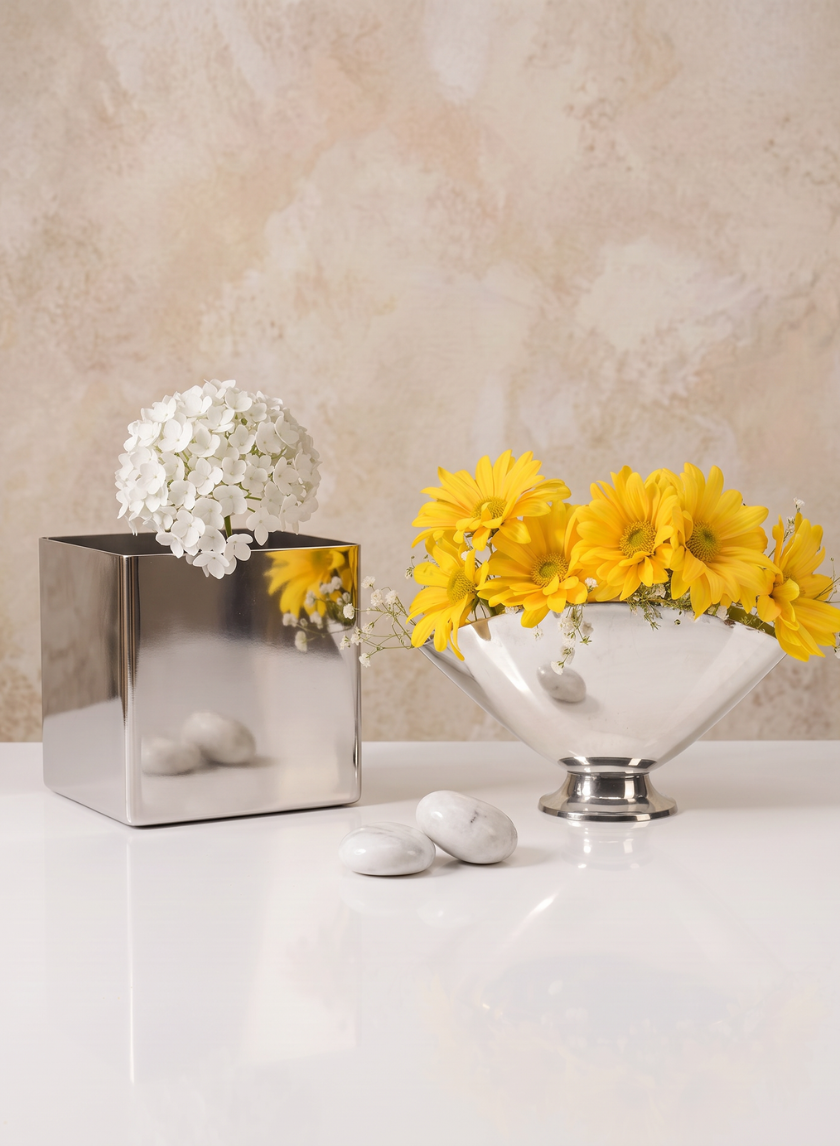 A 5in Cube Polish Stainless Steel Vase with white flowers and a shiny bowl filled with yellow daisies sit on a white surface. Two white stones are placed in front of the bowl, all arranged against a light textured background.
