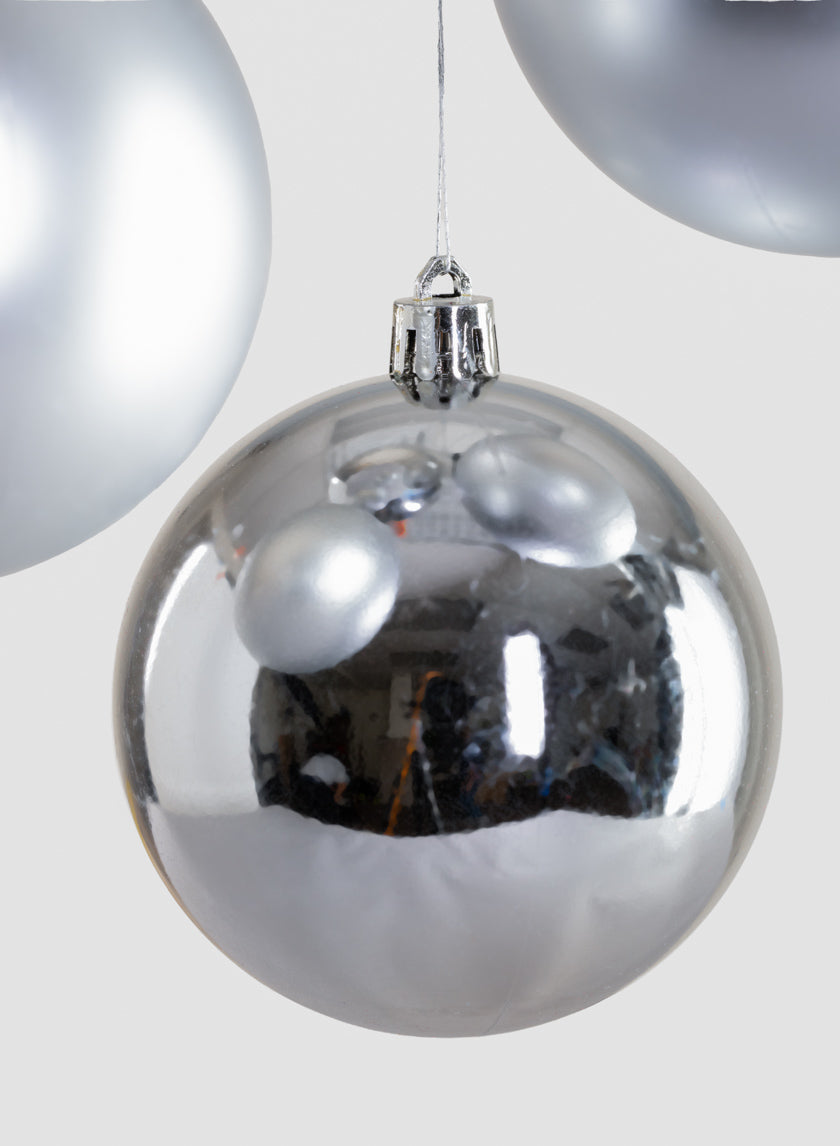 A close-up of the 3in Round Matte & Shiny Silver Ornament Ball hanging on a string, its shiny surface reflecting light, with other similar ornaments partially visible in the background.