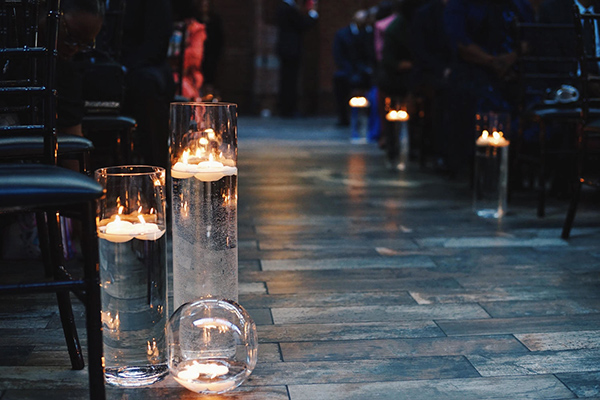 A ceremony aisle with groups of floating candles in three vessels: two glass cylinder vases and a ball votive holder.