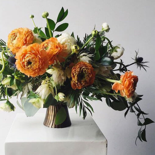 antique gold pedestal bowl with orange ranunculus thistle and other greenery