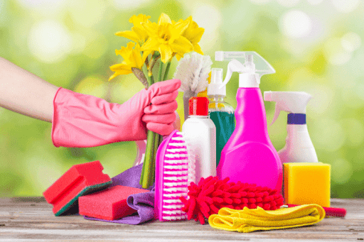 A woman's hand holds a cleaning product and supplies, with a silk flower in the background.