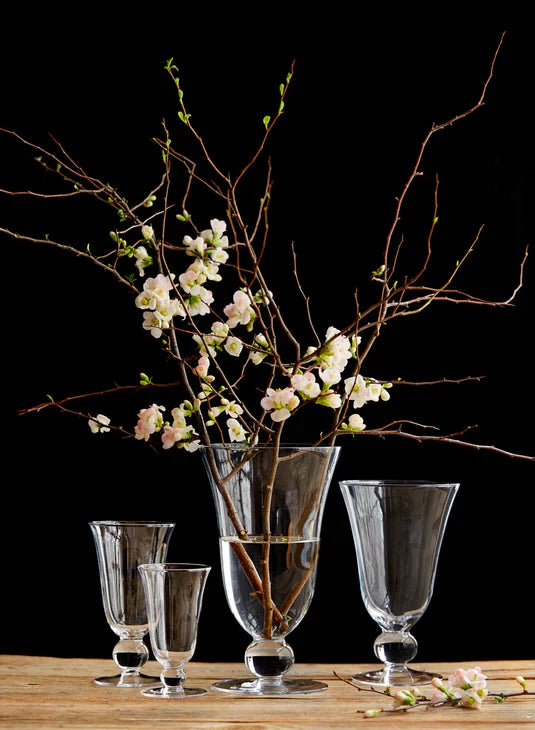 A clear vase with water holds six vibrant orange-red calla lilies, set on a wooden surface against a neutral, textured background.
