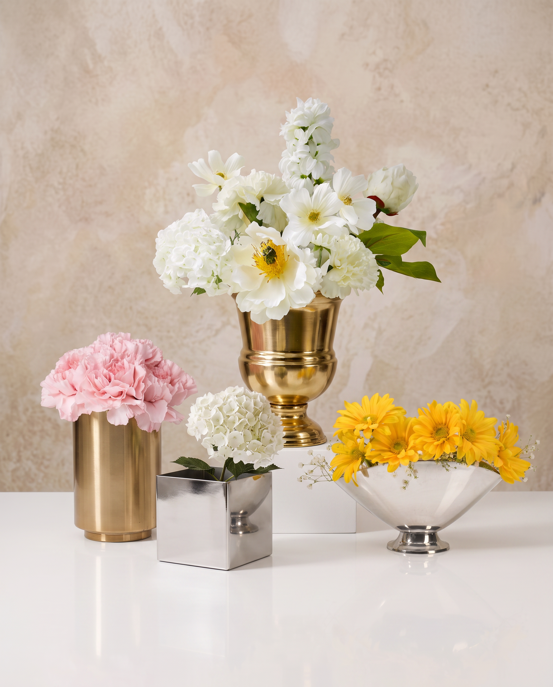 Three gold vases sit on a wooden surface; two hold white flowers with green leaves, while the third vase is empty. The background is a beige textured wall.