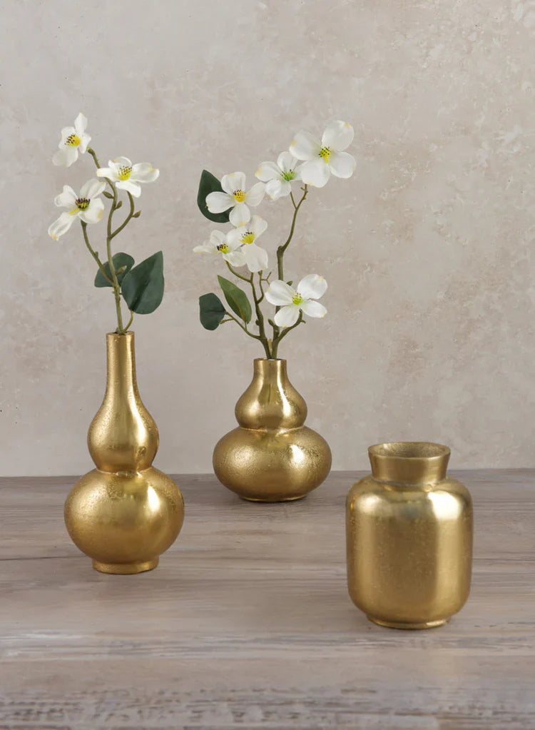 Three gold vases sit on a wooden surface; two hold white flowers with green leaves, while the third vase is empty. The background is a beige textured wall.