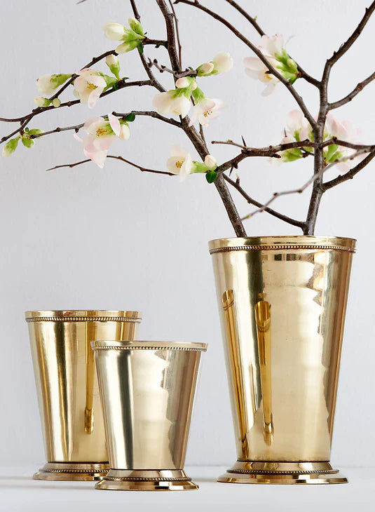 Three gold vases sit on a wooden surface; two hold white flowers with green leaves, while the third vase is empty. The background is a beige textured wall.