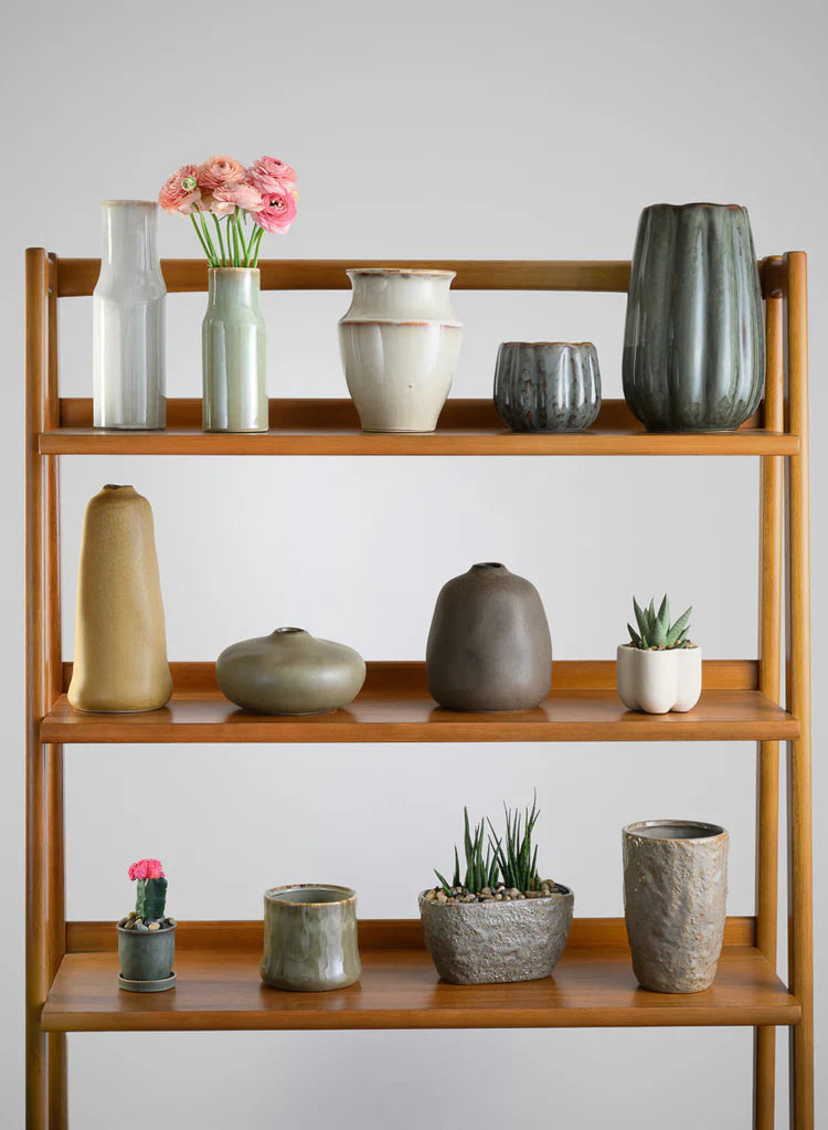 A wooden shelf displays ceramic vases, bowls, and pots in neutral tones. Some hold flowers or small succulents; others are empty. The background is plain and light gray.