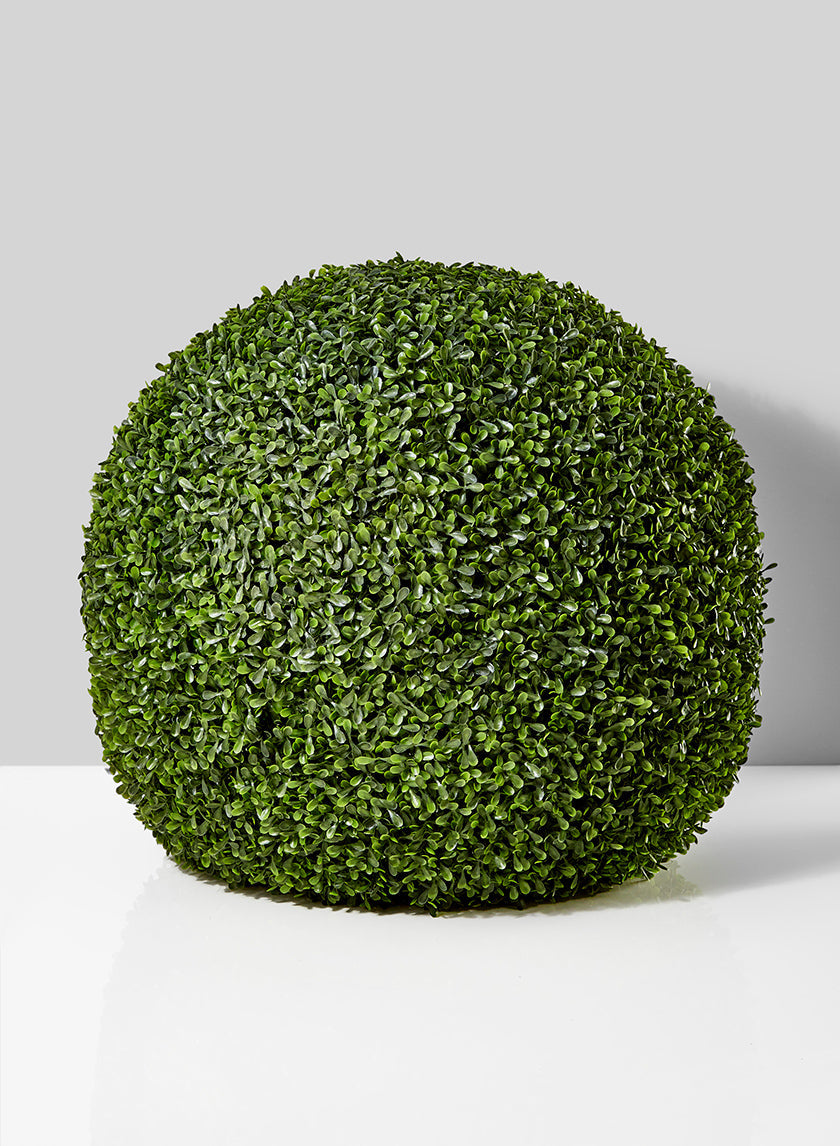 A round, neatly trimmed artificial boxwood bush sits on a white surface against a light gray background.
