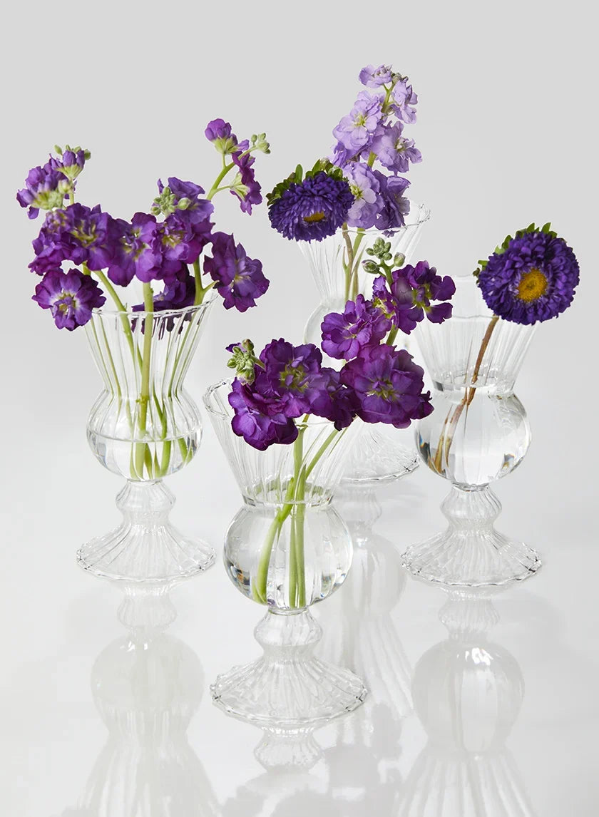 A green glass vase holds three pale green hydrangea blooms with large green leaves, set on a wooden surface against a neutral beige background.