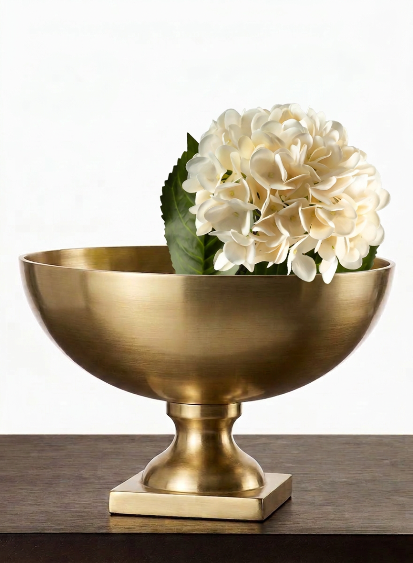 A single cream-colored hydrangea with green leaves is displayed in a 12in Wide Amalfi Gold Footed Centerpiece Compote Metal Bowl, placed on a dark brown surface against a white background.