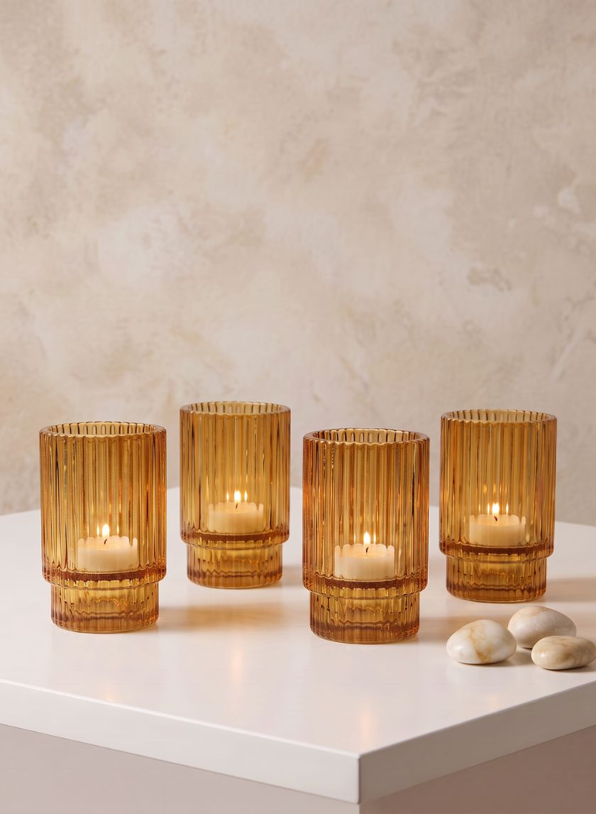 A set of four 3.75in tall amber pleated glass votive holders are displayed on a white surface, each holding a lit tealight candle. They are arranged next to three smooth white stones, against a light textured background.