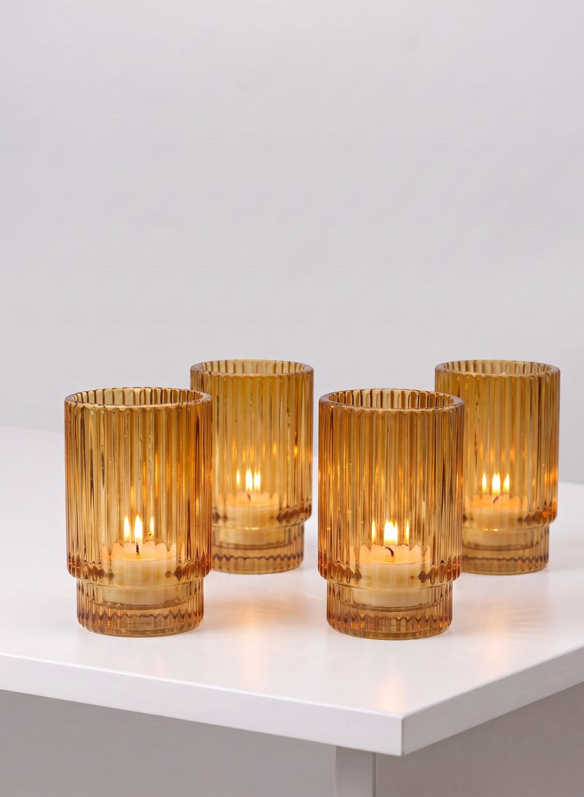 Set of four 3.75in tall amber pleated glass votive holders are arranged on a white surface against a plain light gray background, each holding a lit candle inside.