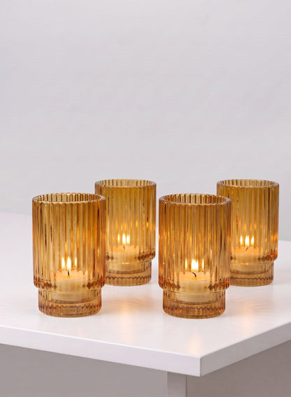 Set of four 3.75in tall amber pleated glass votive holders are arranged on a white surface against a plain light gray background, each holding a lit candle inside.