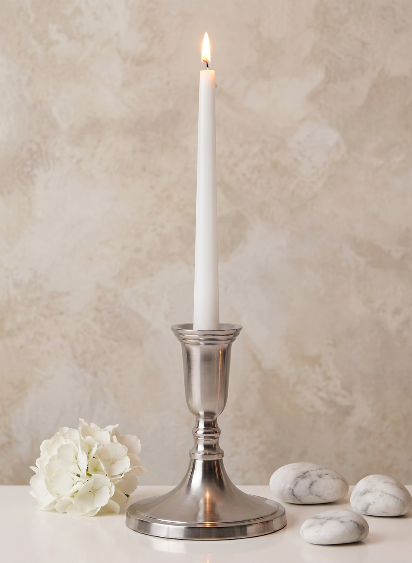 A lit white taper candle is displayed in the 5.5in Tall Nickel Silver Candlestick, placed on a white surface alongside a white flower and three marble stones, set against a beige textured background.