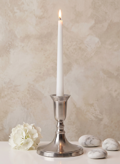 A lit white taper candle is displayed in the 5.5in Tall Nickel Silver Candlestick, placed on a white surface alongside a white flower and three marble stones, set against a beige textured background.