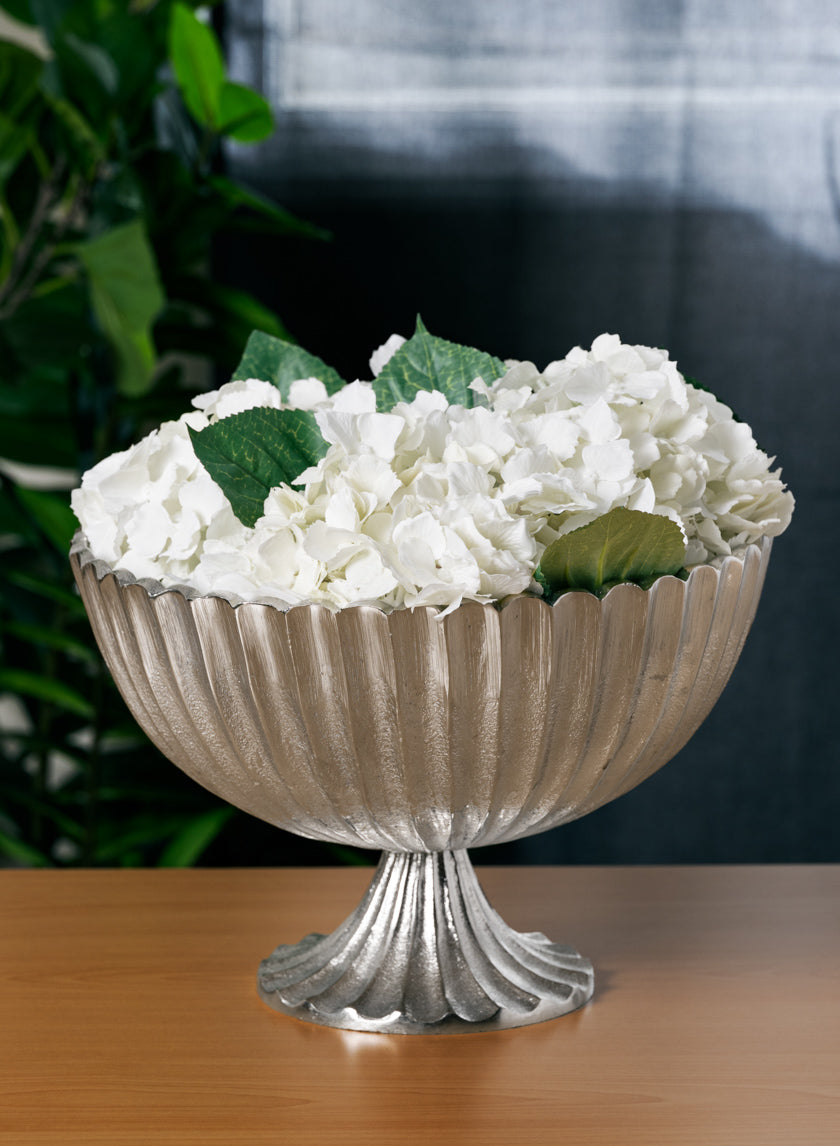 A 12in Palermo Brushed Silver Scallop Compote Metal Bowl, filled with white hydrangeas and green leaves, sits on a wooden surface with a green plant and dark curtain in the background.