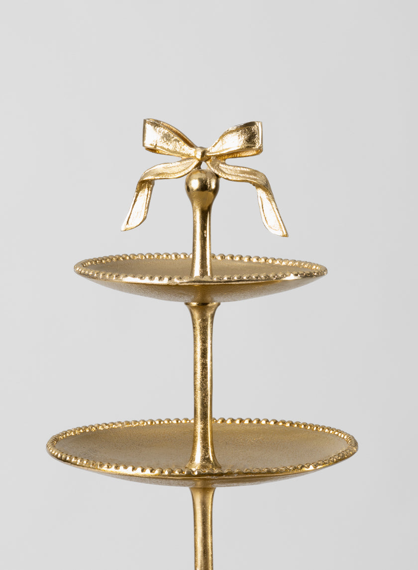 The 21in Tall Atelier 3 Tier Gold Display Stand features three round plates with beaded edges and a decorative bow on top, set against a plain light background.