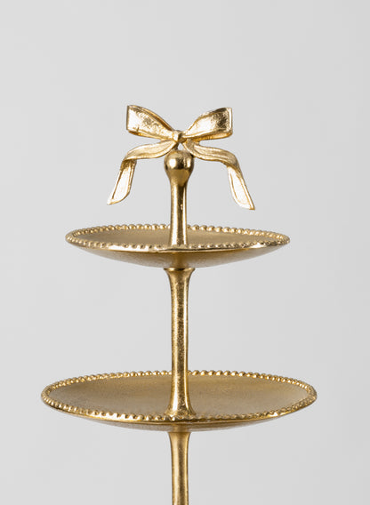 The 21in Tall Atelier 3 Tier Gold Display Stand features three round plates with beaded edges and a decorative bow on top, set against a plain light background.