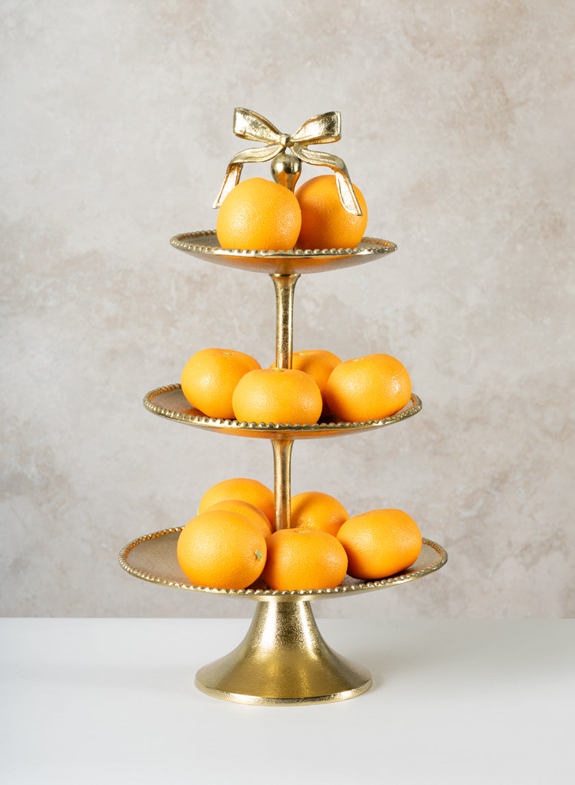 The 21in Tall Atelier 3 Tier Gold Display Stand, topped with a decorative bow, elegantly holds multiple whole oranges on each tier against a light textured background.