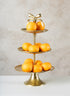 The 21in Tall Atelier 3 Tier Gold Display Stand, topped with a decorative bow, elegantly holds multiple whole oranges on each tier against a light textured background.