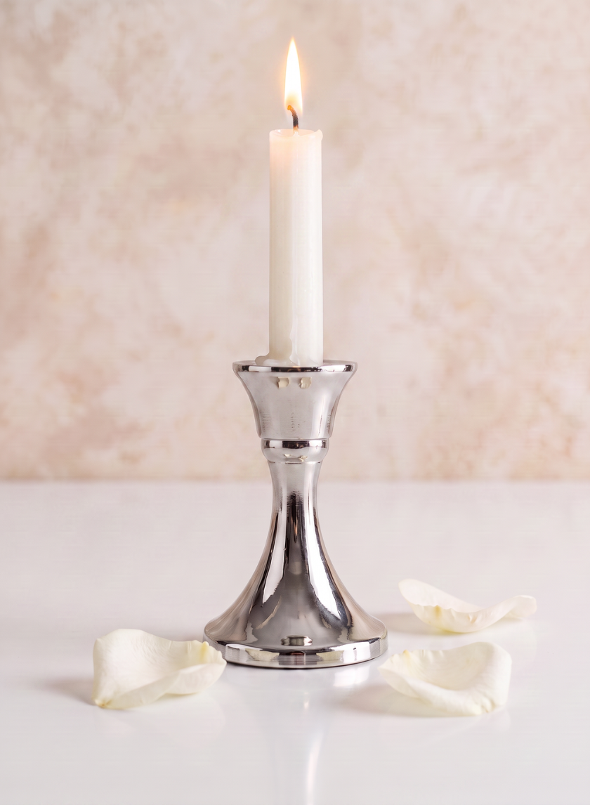 A white candle is displayed in a 4.5in Tall Silver Metal Candlestick on a white surface, surrounded by scattered white rose petals, set against a soft beige background.