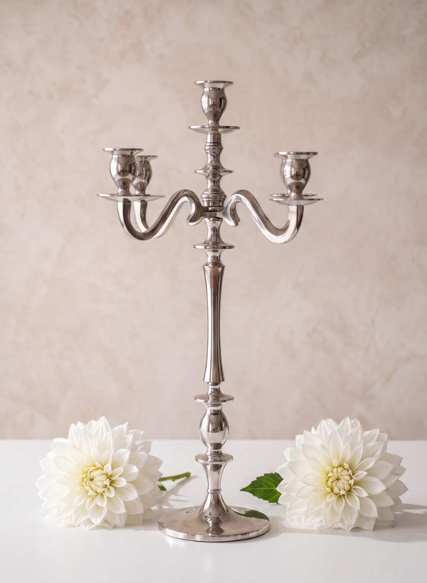 A 25.75in Tall Silver Metal Candelabra with five arms is displayed on a white surface, flanked by two large white flowers, against a soft beige background.