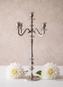 A 25.75in Tall Silver Metal Candelabra with five arms is displayed on a white surface, flanked by two large white flowers, against a soft beige background.