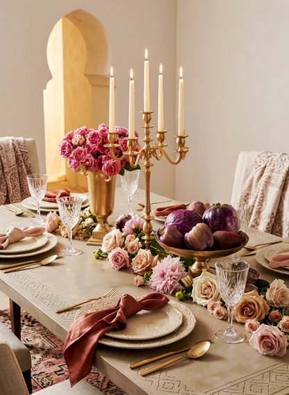 An elegant dining table features gold utensils, crystal glasses, and plates with pink napkins. Gold Metal Candle Holders, pink roses, and purple vegetables create a luxurious, romantic centerpiece for the event decor.