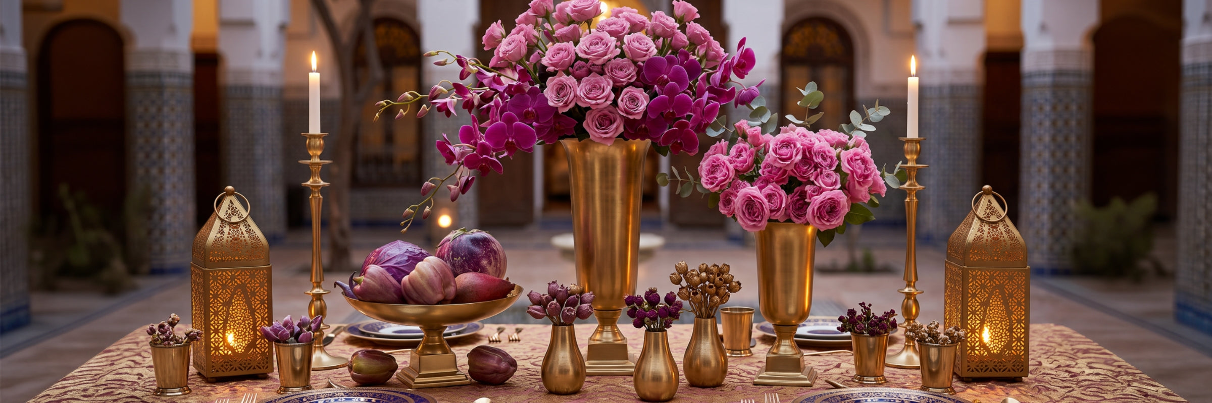 A beautifully arranged Moroccan-style table setting featuring large gold vases filled with pink and purple roses, ornate gold lanterns, lit candles, and a bowl of fruit, set against a blurred background of an arched courtyard.