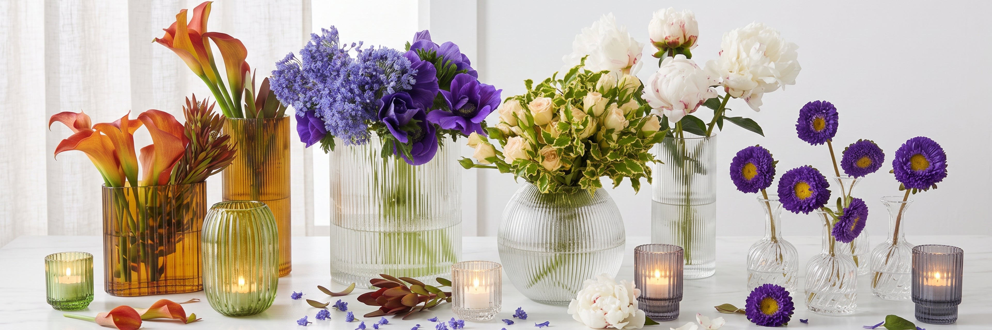 A styled tabletop arrangement of multiple flower arrangements in clear ribbed glass vases and amber glass containers, with lit tealight candles and scattered flower petals on a white surface.