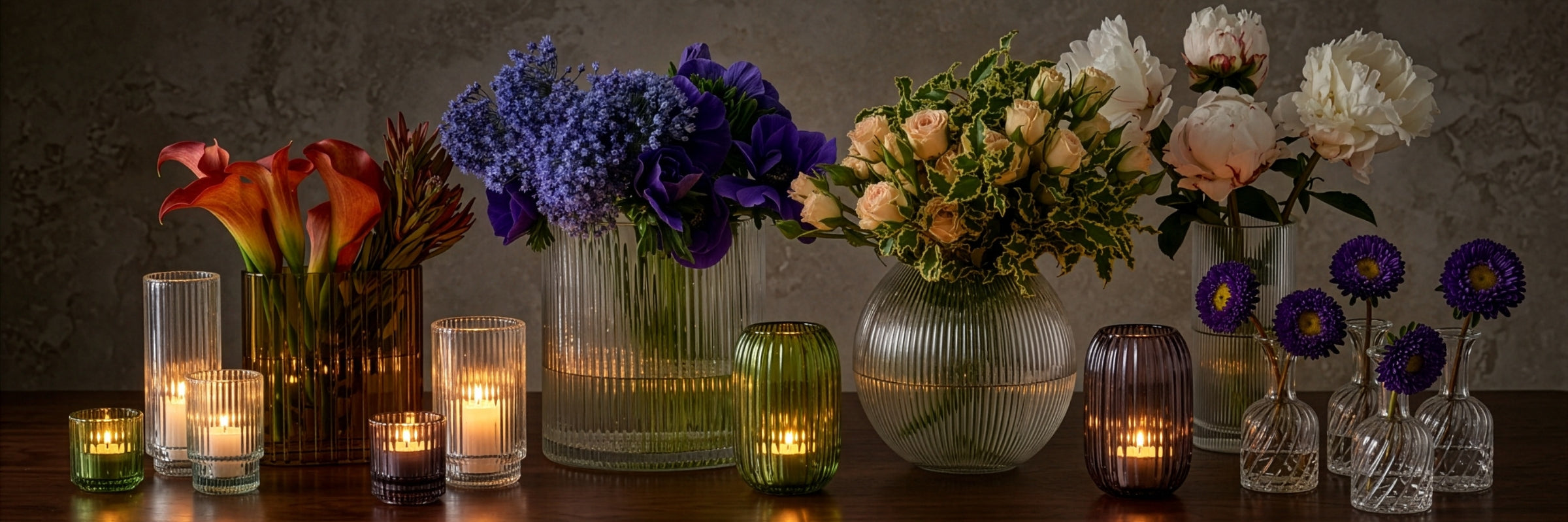 A styled arrangement of multiple glass vases and candle holders on a dark table, featuring floral arrangements in purple, cream, and orange-red tones, with lit candles in clear and tinted glass against a textured gray wall.