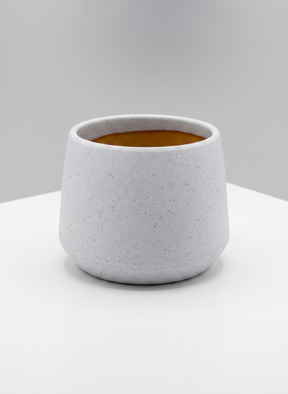 A 4in Tall Florist Choice Matte White Bell Cachepot, shown here with a round silhouette and speckled ceramic finish, features a wide opening and sits on a white surface against a light gray background. The interior of the cachepot displays a warm brown hue.