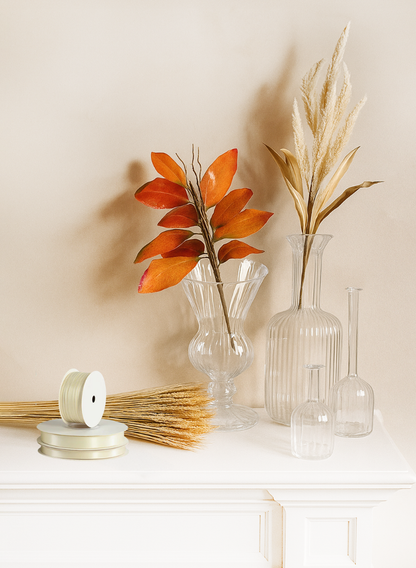 A 35in Long Beige Amaranth Reed (Case of 12) is arranged in glass vases with dried pampas grass and orange leaves on a white surface, complemented by wheat bundles and cream ribbon for minimalist, autumn-themed decor.