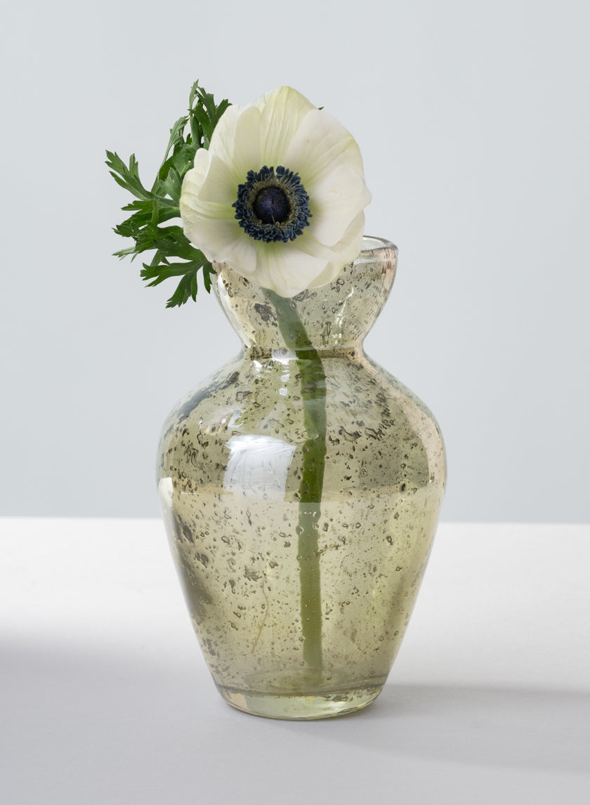 A single white anemone with a dark center and green leaves sits in a 6in Tall Jacques Green Vintage Bud Glass Vase, displayed on a light surface against a plain gray background.