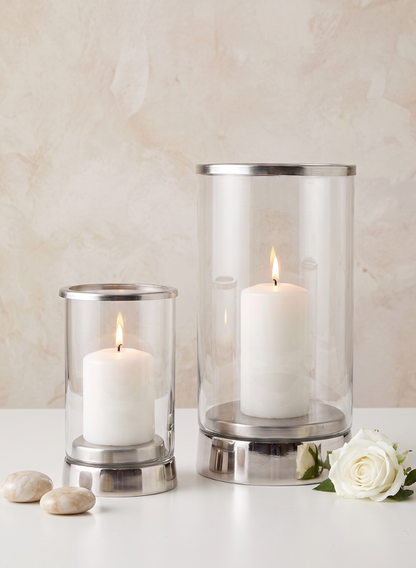 Two 7in Tall Silver &amp; Glass Candle Hurricanes, each holding a white pillar candle, are set on a white surface. Next to them lie two smooth stones and a single white rose, with a softly textured light wall in the background.