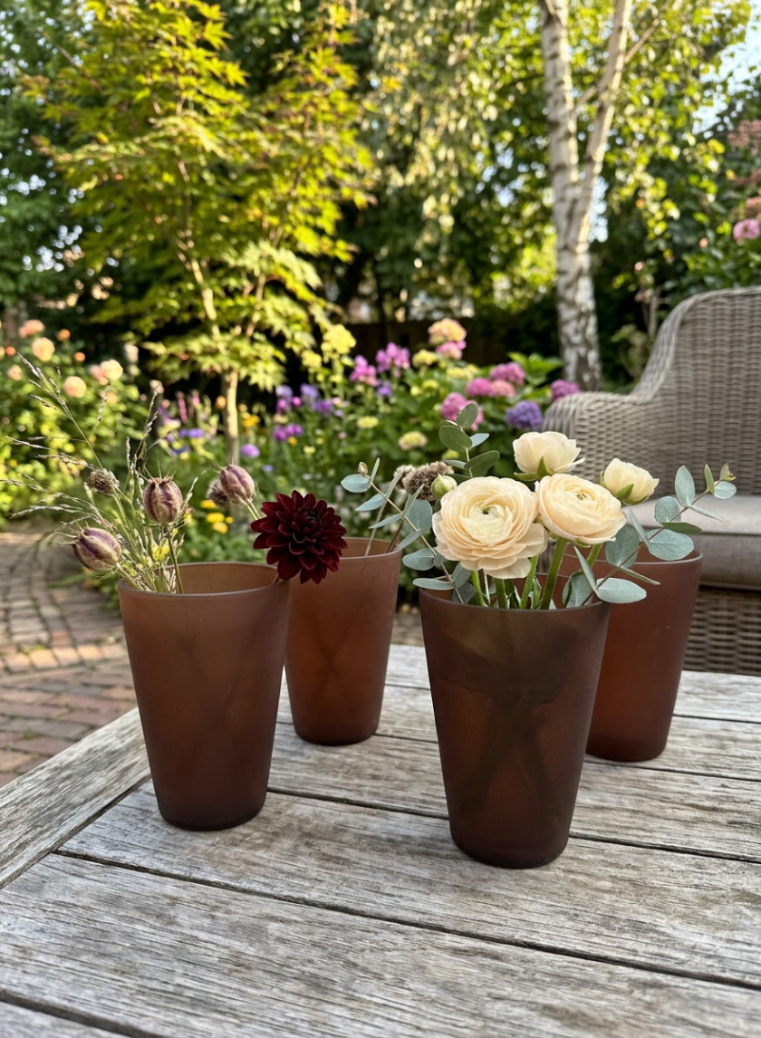 Four 4in Tall Terracotta-Look Deep Brown Glass Vases, set of 4, with mixed flowers like pale yellow roses and a dark red bloom, are displayed on a wooden table as rustic decor in a sunlit garden featuring trees and wicker furniture.