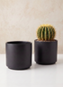 A round cactus with yellow spines sits in one of the 5.25in Tall Florist Choice Matte Black Cylinders, filled with small pebbles, next to an identical empty cylinder from the set of two on a white surface against a beige wall.