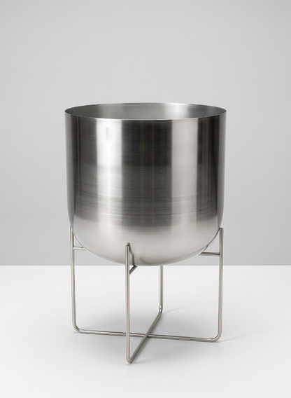 A 23in Tall Silver Planter With Stand, featuring a large brushed stainless steel container supported by a minimalistic metal base with four slender legs that cross at the bottom, is displayed against a plain light gray background.
