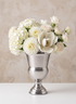 An 11.25in Tall Silver Urn Metal Vase filled with white hydrangeas, dahlias, magnolia, and roses sits on a white surface against a light beige, textured background.