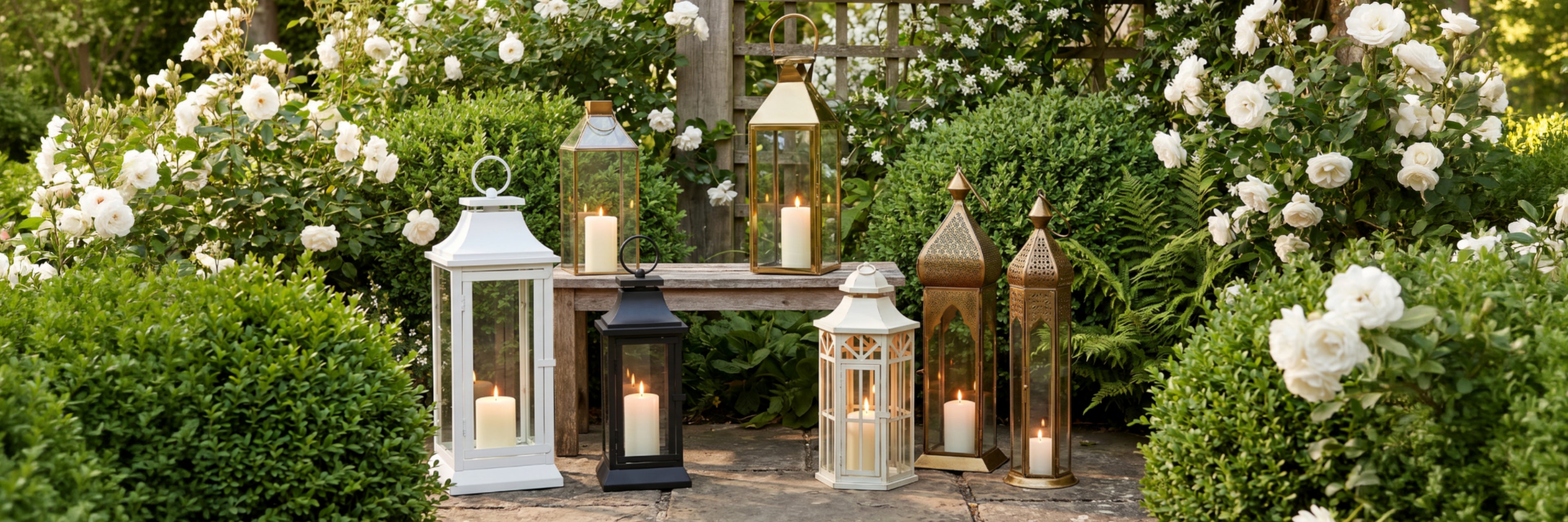 A set of six decorative outdoor lanterns with glass panels and lit candles, arranged on a stone path in a garden surrounded by white roses and green foliage.