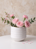 Pink roses and carnations with greenery arranged in a 5in Clay White Porcelain Cylinder with Gold Edging, set on a white surface with a few scattered petals, against a beige textured background.