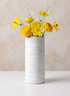 A 4in x 10in Clay White Porcelain Cylinder with Gold Edging, filled with bright yellow flowers, sits on a white surface against a light beige background.