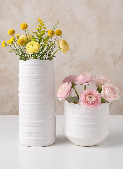 Two white porcelain vases sit on a white surface. The taller vase, a 5in Clay White Porcelain Cylinder with Gold Edging, holds yellow and green flowers, while the shorter vase displays pink blooms. The background is light beige with a subtle textured pattern.