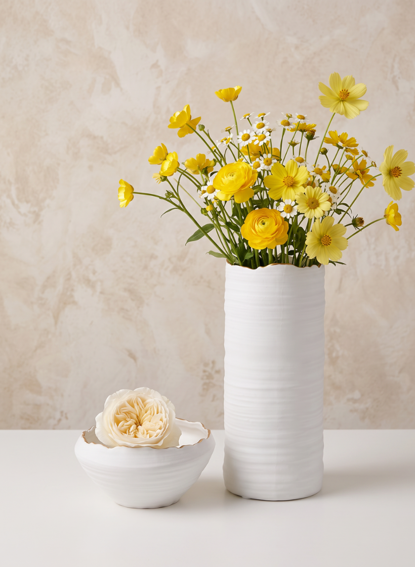 A tall white vase with yellow wildflowers and a 6in Clay White Porcelain Bowl with Gold Edging holding a pale cream rose, set on a white surface against a beige textured background.